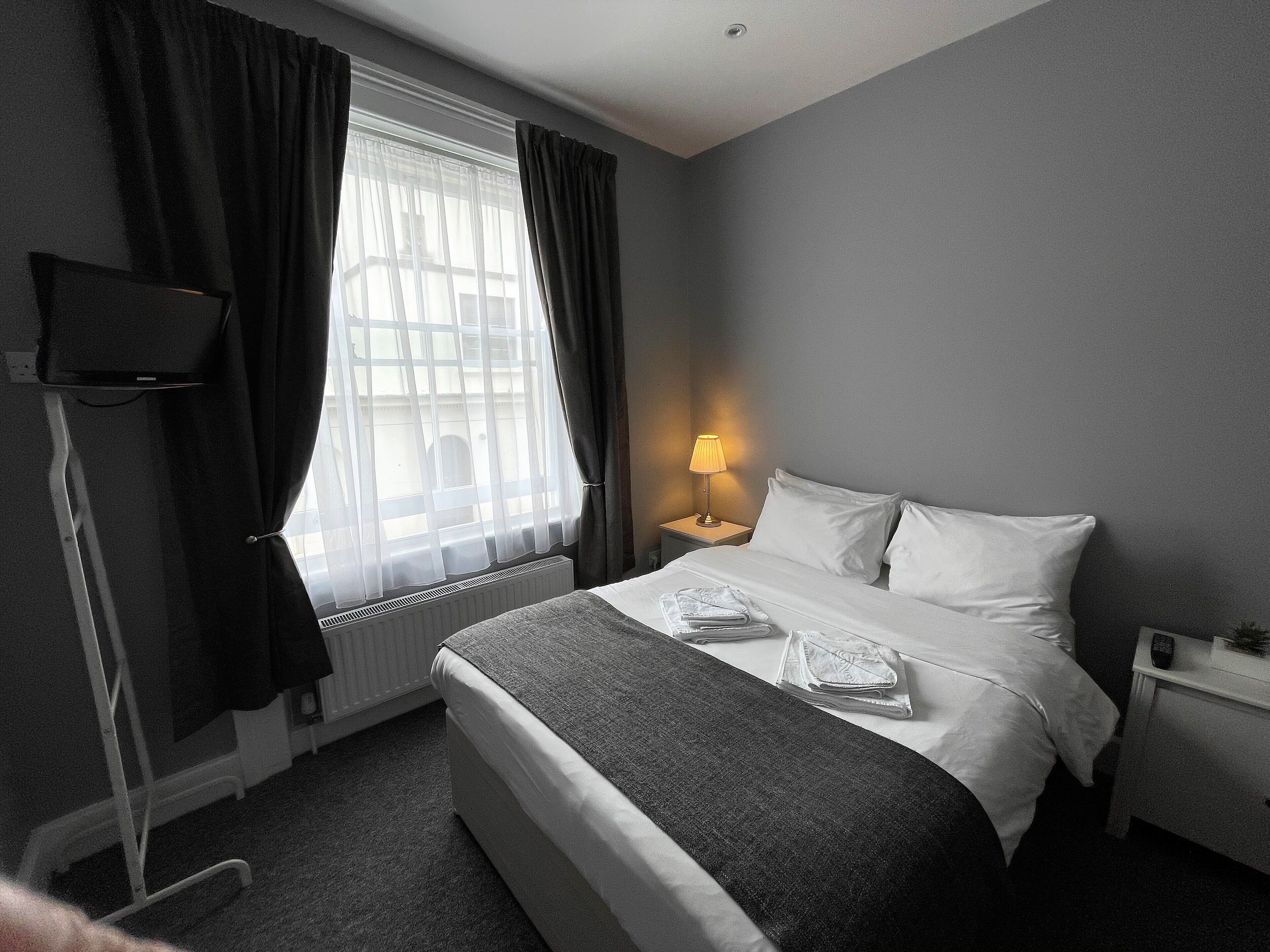 A hotel room with a bed, a bedside lamp, a TV on a stand, and a window with curtains.