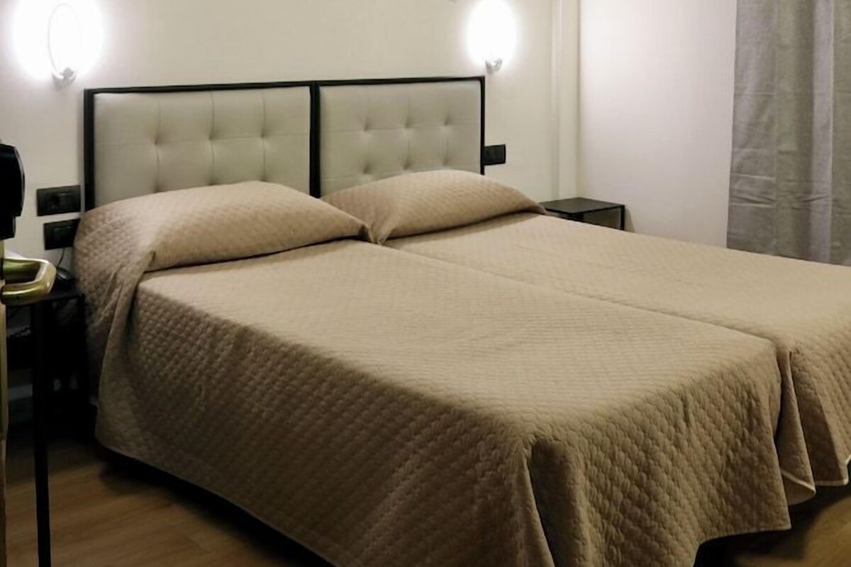 A bedroom with a double bed, two bedside tables, a wall-mounted light fixture, and a curtain.