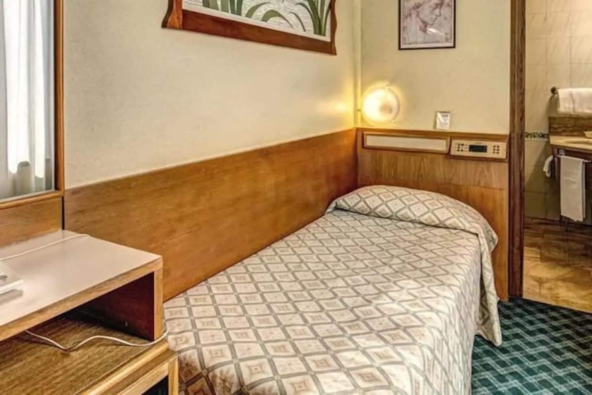 Ein Einzelbett mit gemustertem Bettbezug, ein Nachttisch mit Lampe und ein kleiner Schreibtisch mit Stuhl.