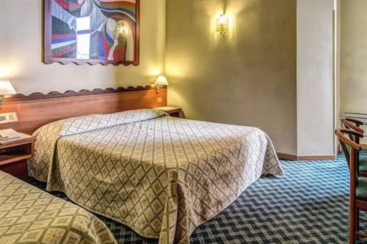 Ein Hotelzimmer mit zwei Betten, einem Holzkopfende, Nachttischlampen, einem Nachttisch, einem Wandbild und einem gemusterten Teppich.