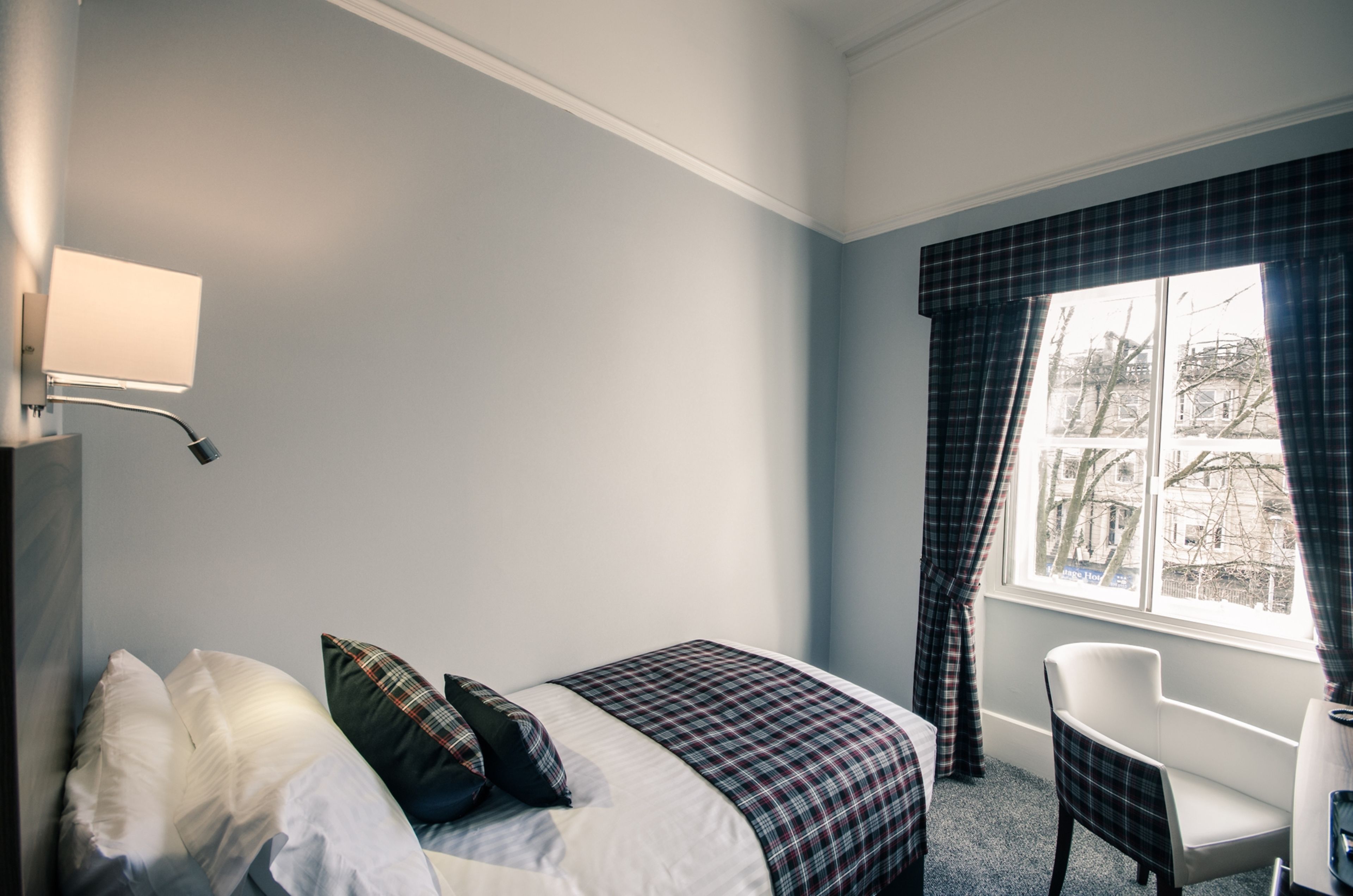 A bedroom with a bed, pillows, a chair, a window with curtains, and a wall-mounted lamp.