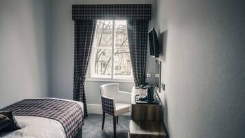 Habitación de hotel con cama, escritorio, silla, ventana con cortinas y televisor de pantalla plana montado en la pared.