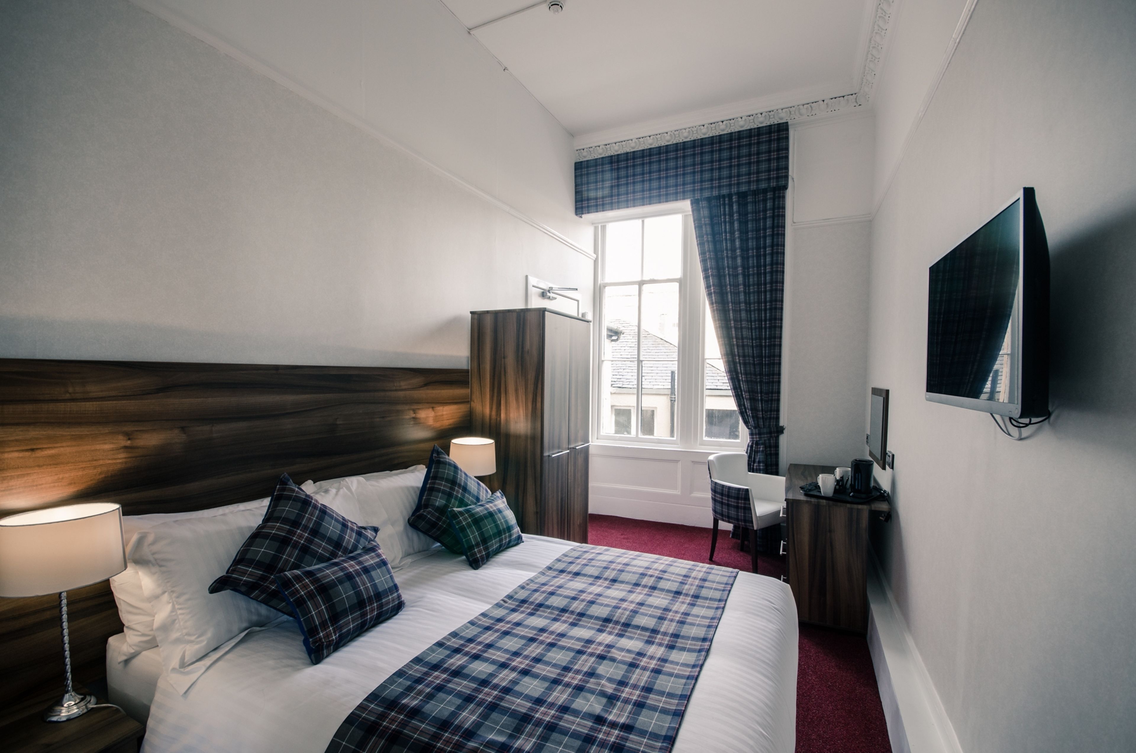 A hotel room with a wooden headboard, a bed with plaid bedding, a bedside lamp, a desk with a chair, a television mounted on the wall, and a window with curtains.