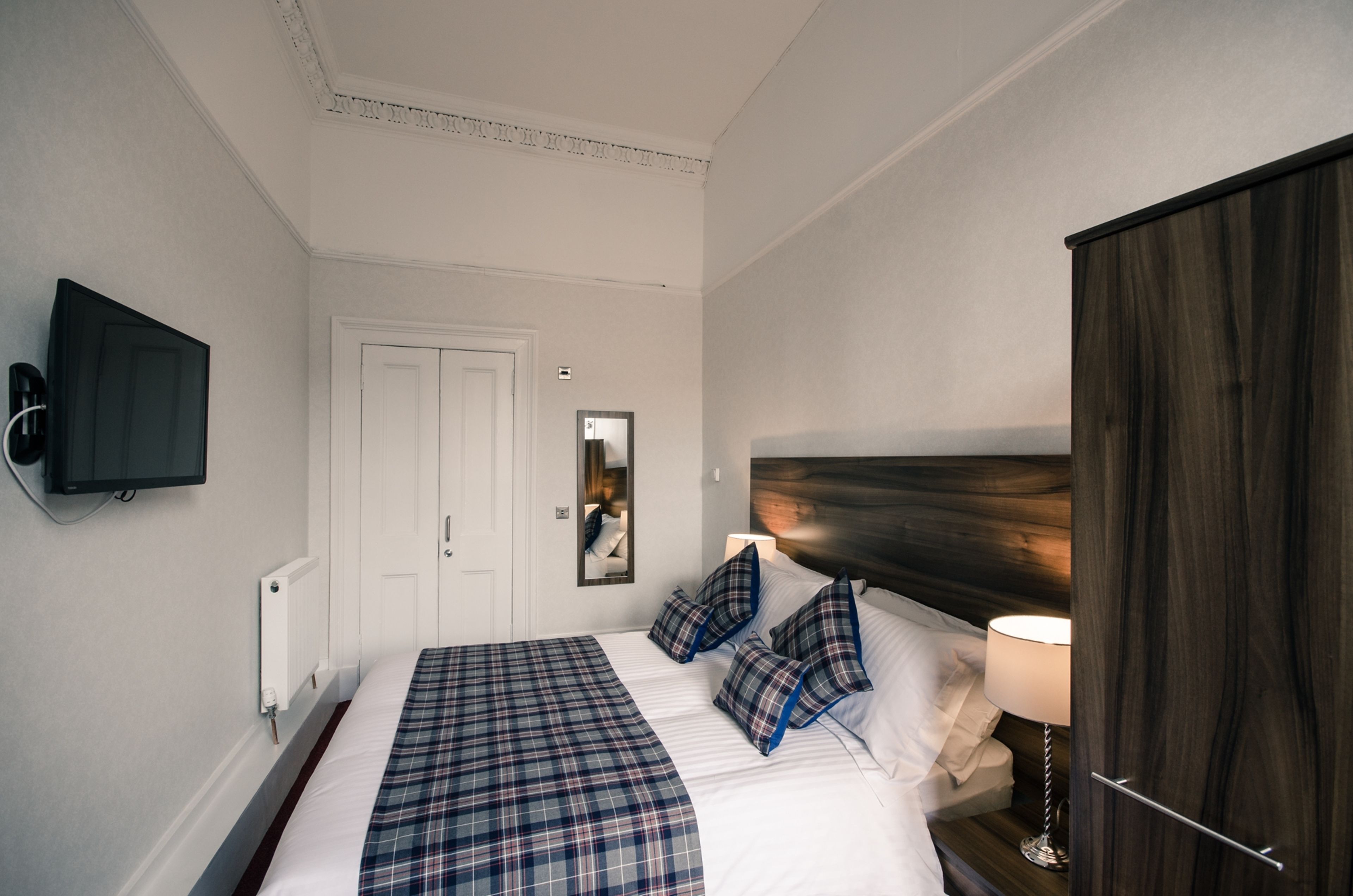 A hotel room with a large bed, a television mounted on the wall, a wooden headboard, and a mirror.