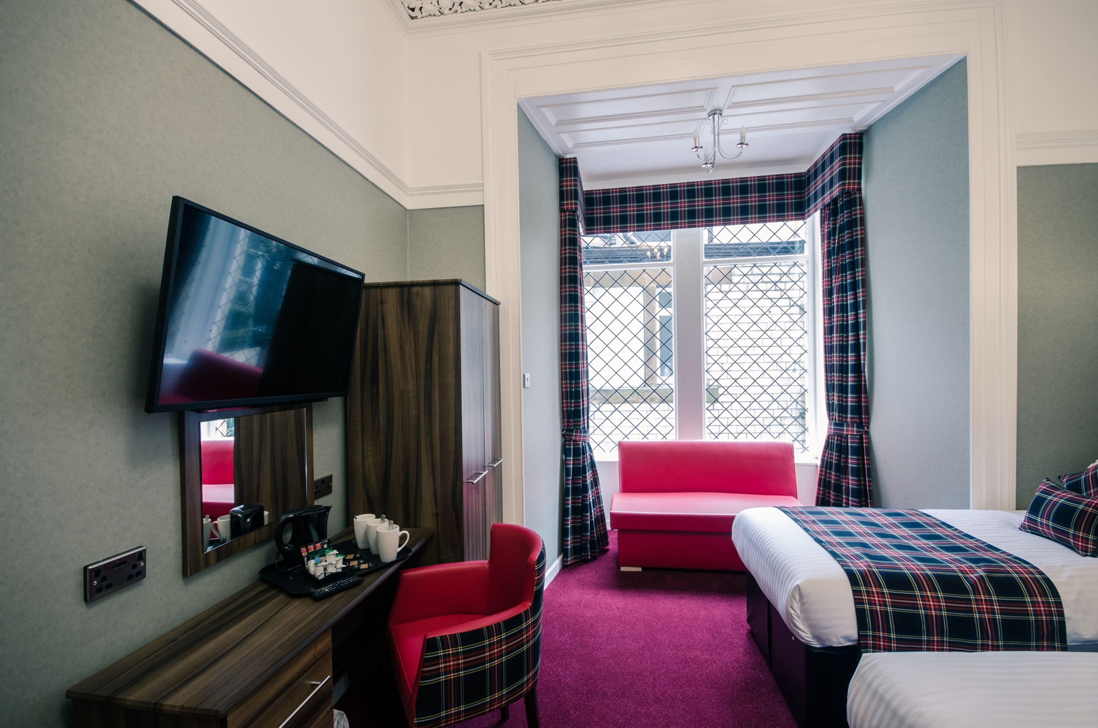 A hotel room with a television, a desk with a coffee maker, a red chair, a bed with plaid bedding, and a window with curtains.