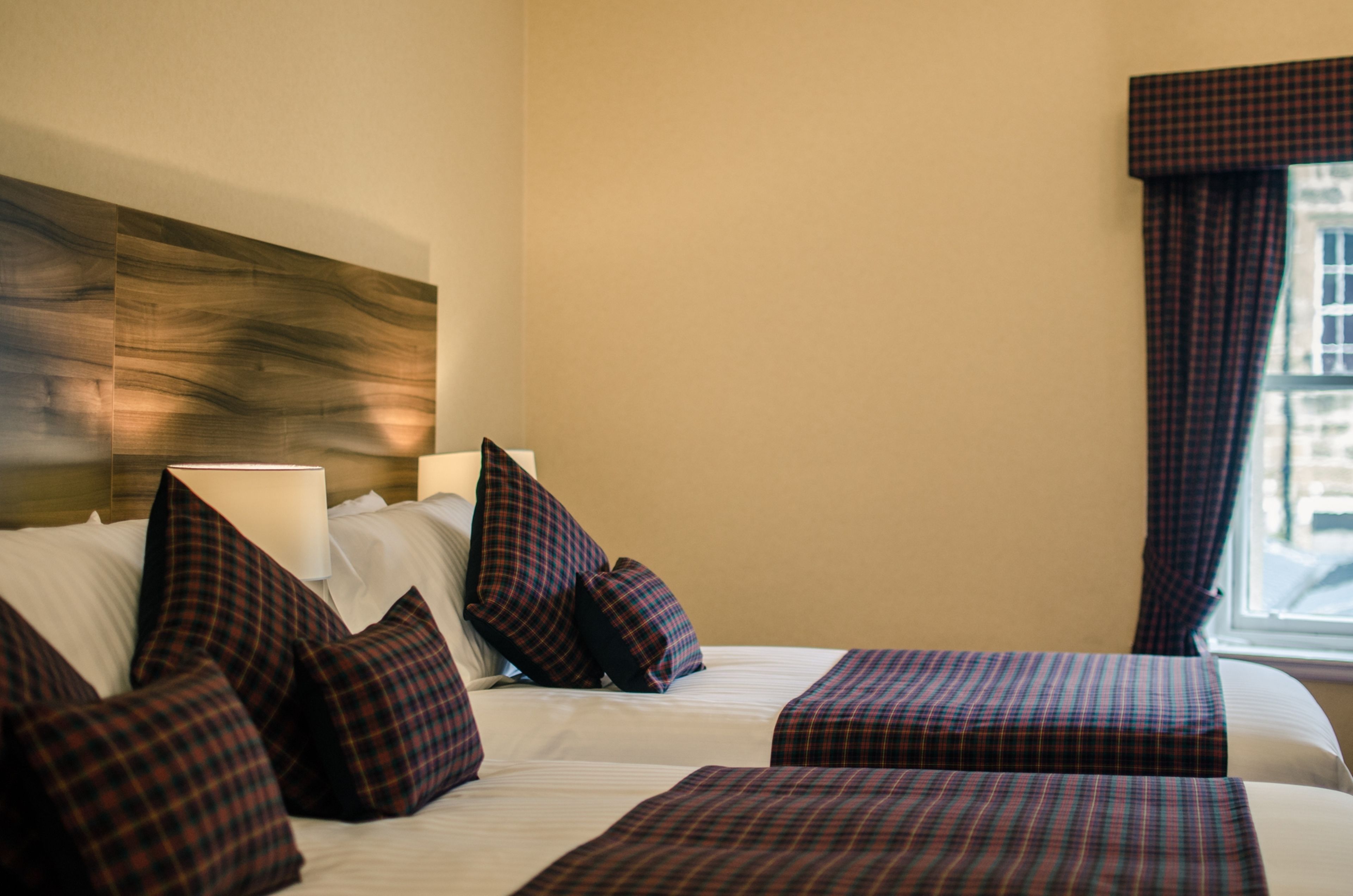 Een hotelkamer met twee bedden, ruitjes beddengoed, een hoofdeinde, een raam met gordijnen en een wandlamp.