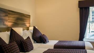 A hotel room with two beds, plaid bedding, a headboard, a window with curtains, and a wall-mounted lamp.