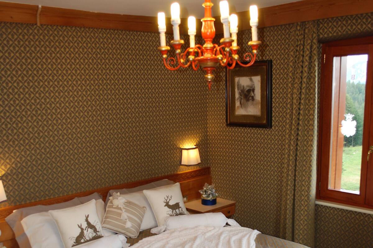 A neatly made bed with patterned bedding, a chandelier, and a nightstand with a lamp.
