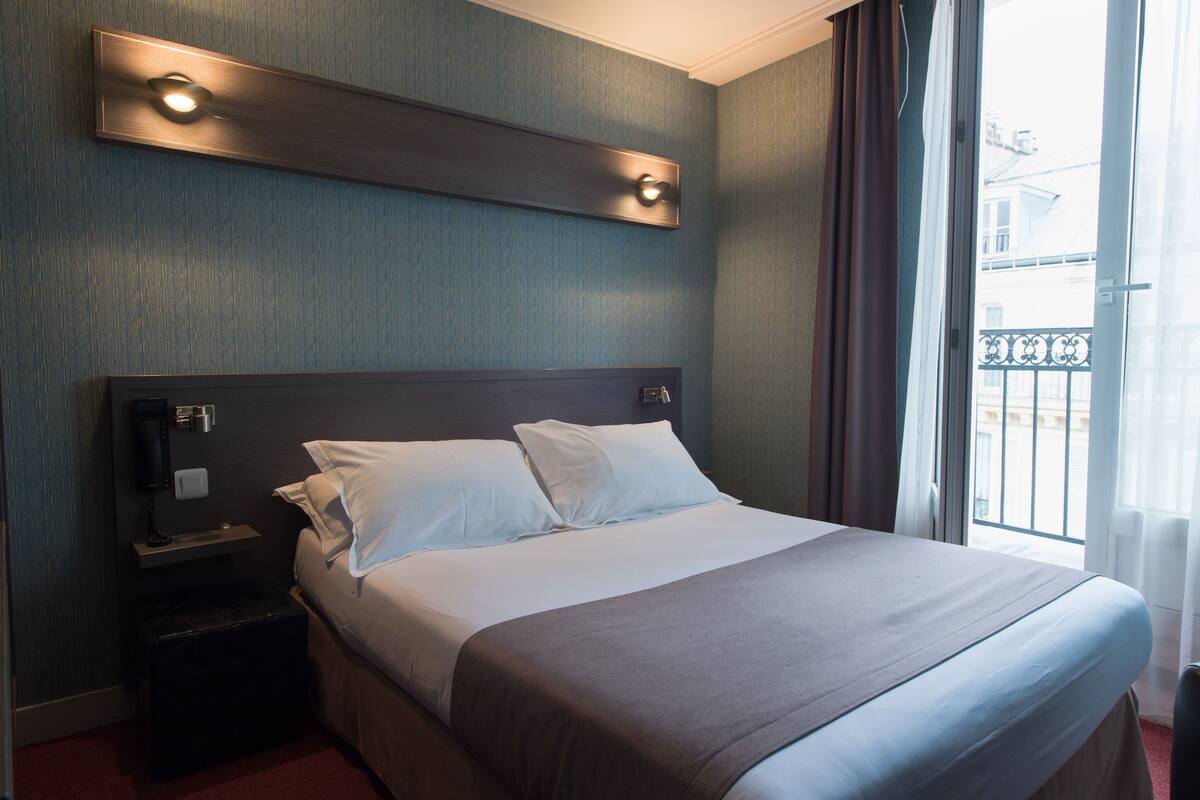 Quarto de hotel com uma cama grande, mesa de cabeceira, luminária na parede e janela com vista para edifícios.