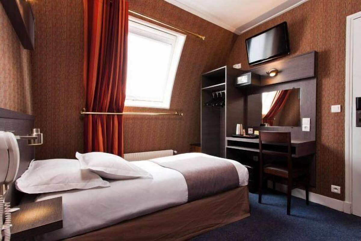 Quarto de hotel com cama, escrivaninha, cadeira, televisão e guarda-roupa.
