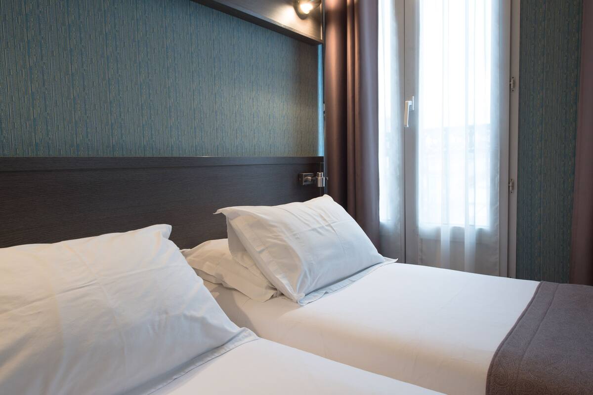 Quarto de hotel com duas camas, cabeceira de madeira, janela com cortinas e parede com textura.