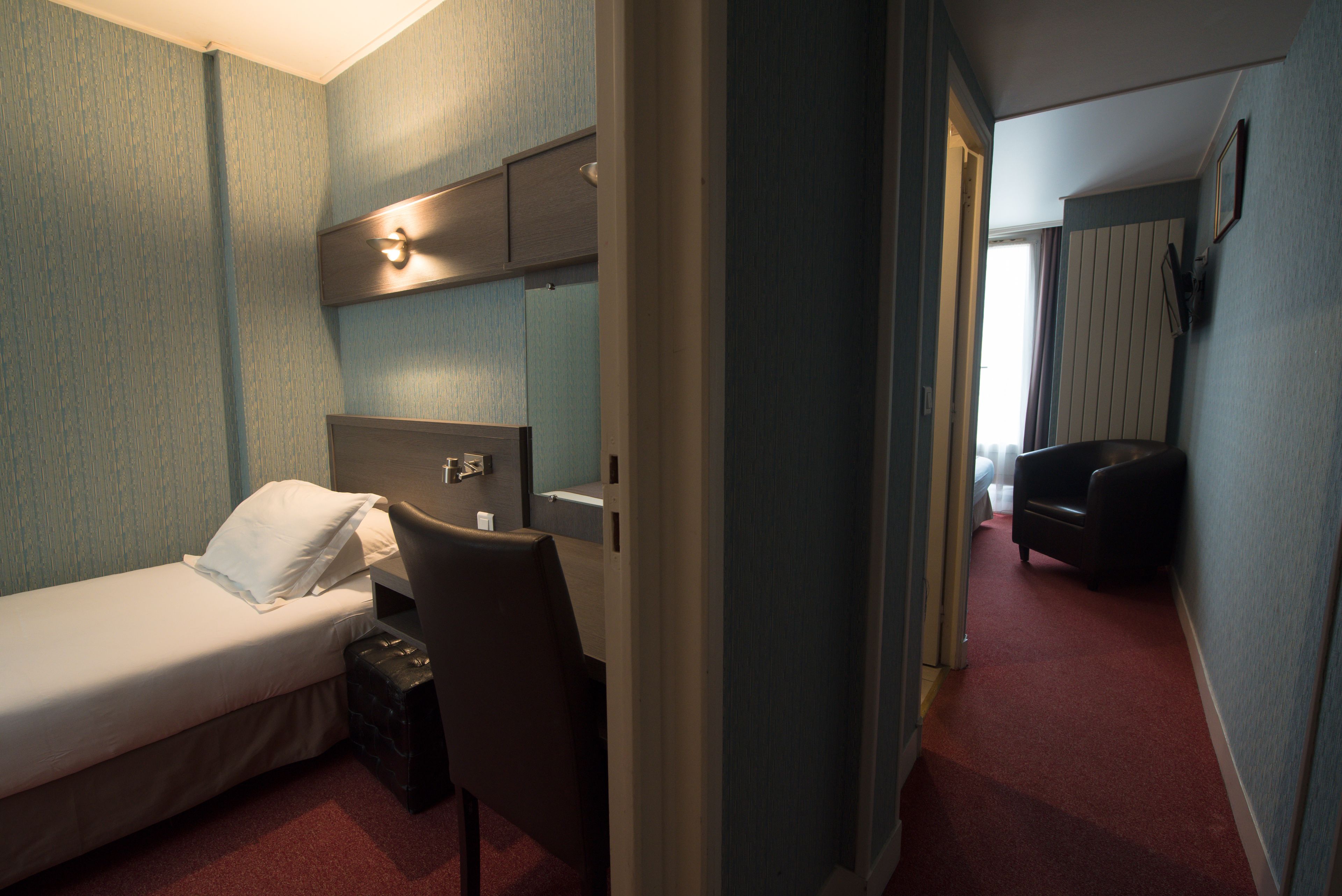 A hotel room with a single bed, a desk, a chair, and a small table. There is a window with curtains, a door leading to another room, and a carpeted floor.
