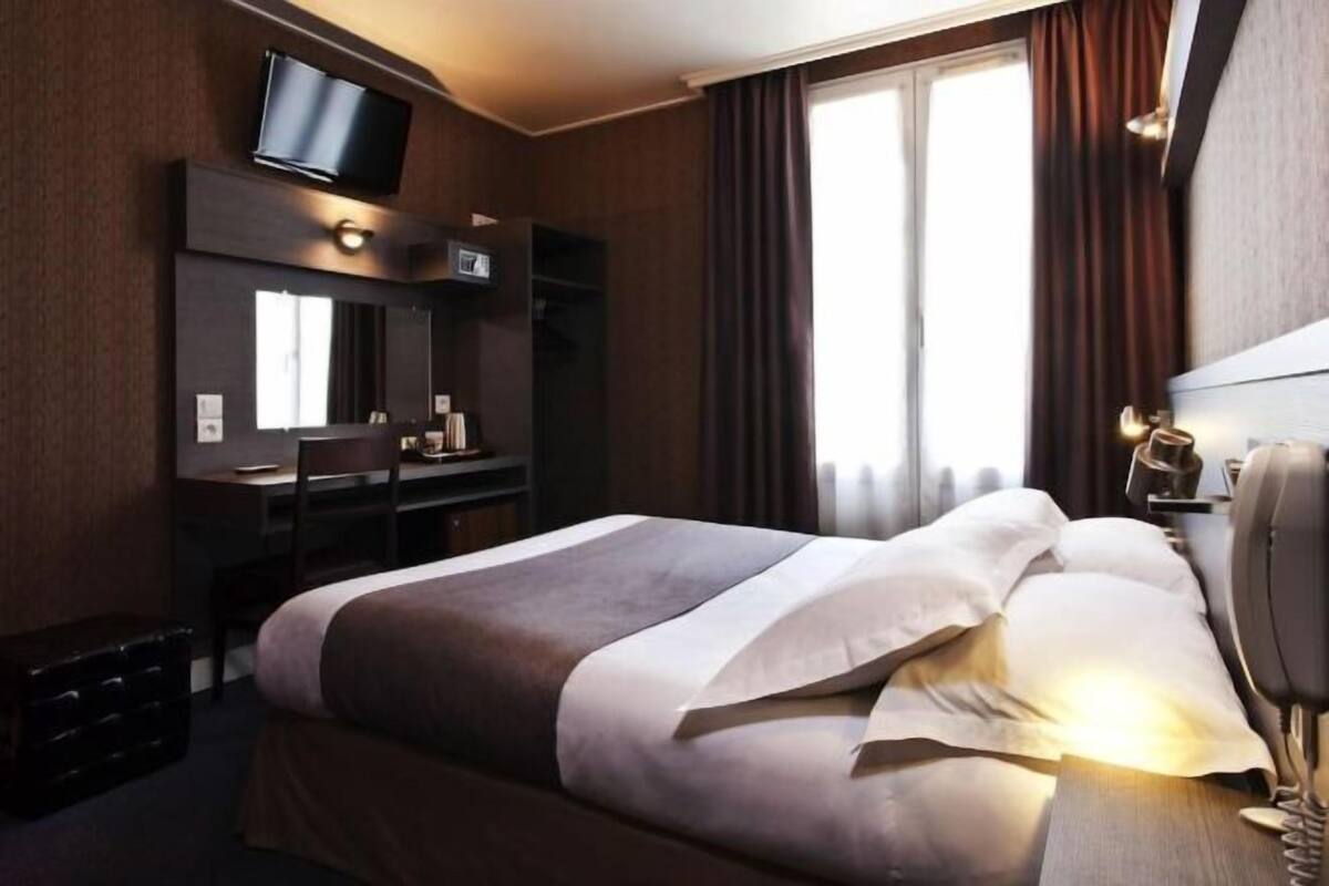 Quarto de hotel com cama, escrivaninha, cadeira, televisão e telefone.
