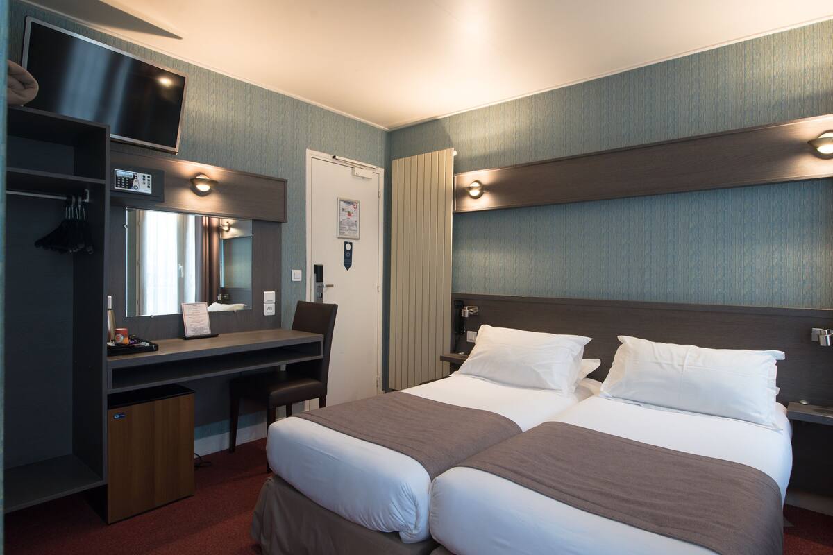 Quarto de hotel com cama, escrivaninha, cadeira e televisão fixada na parede.