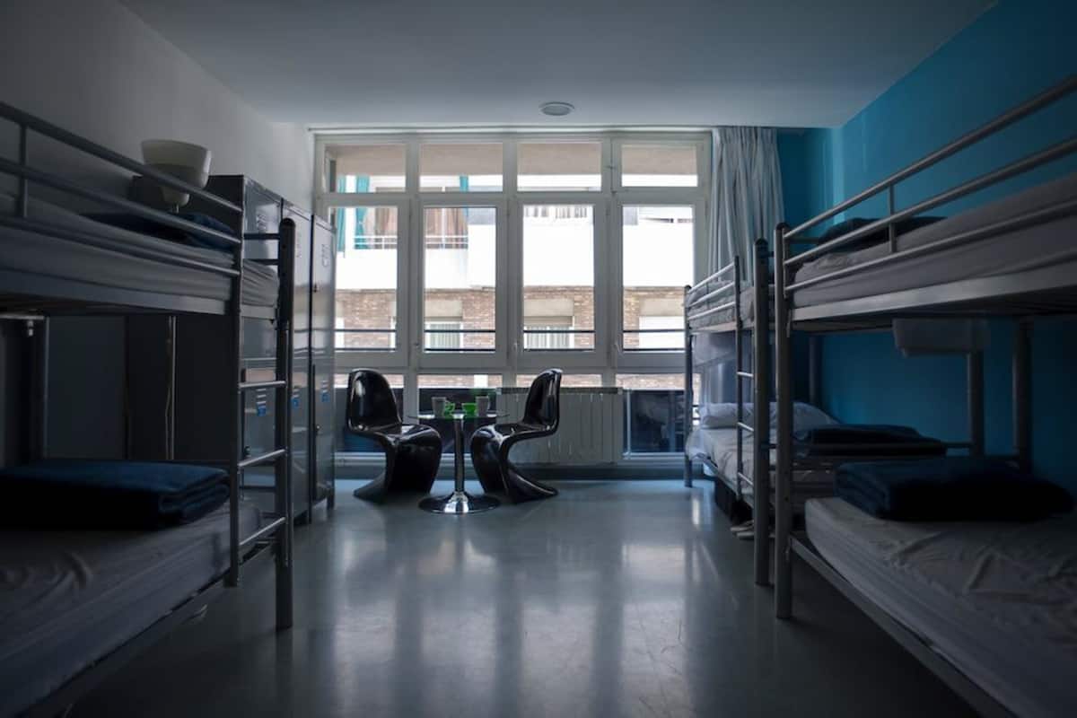 Een studentenkamer met stapelbedden, een tafeltje met stoelen en een raam met gordijnen.