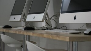 Drei Apple iMac Computer auf einem Holztisch mit weißen Stühlen.