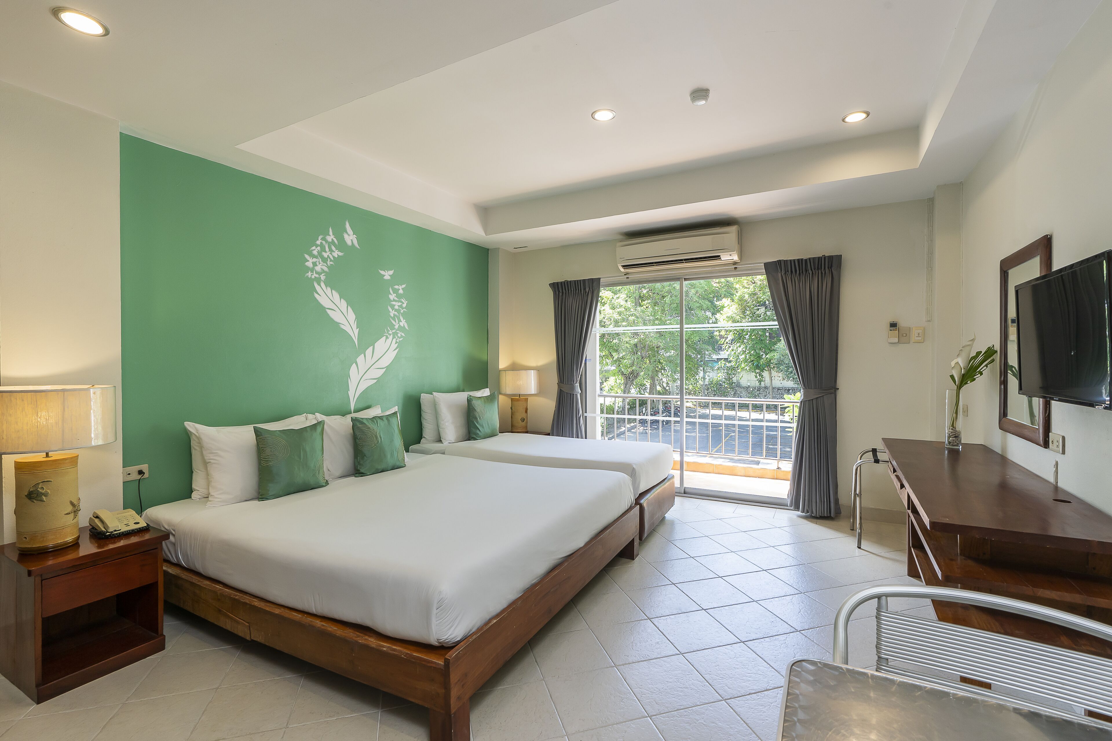 Habitación de hotel con dos camas, televisión, escritorio y balcón con vistas a árboles.