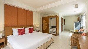 A hotel room with a large bed, a wooden headboard, a wardrobe, a dining area with wicker furniture, and a bathroom with a glass shower.