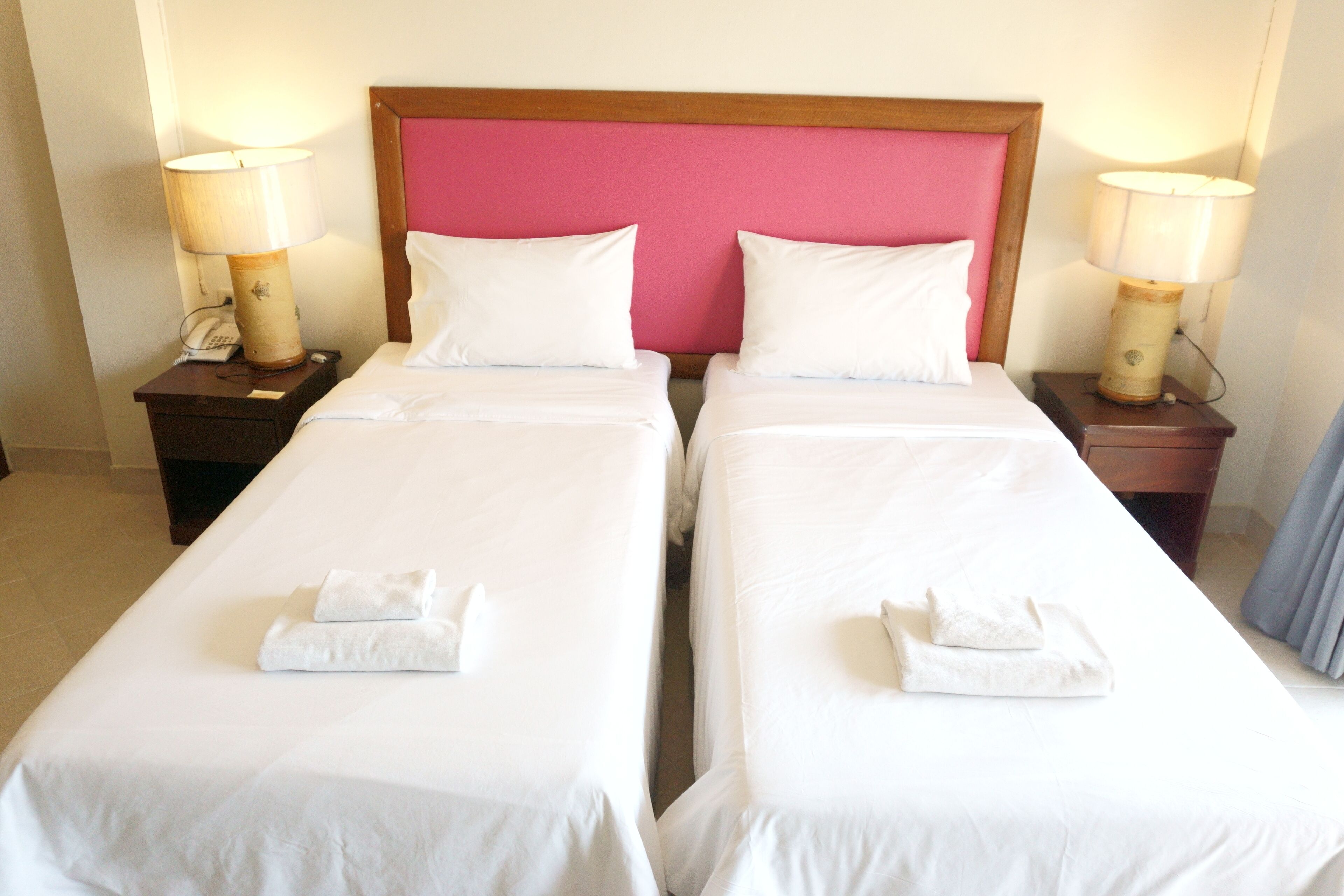 Una habitación de hotel con dos camas individuales, cada una con cabecero de madera, ropa de cama blanca y una lámpara de noche. Hay dos toallas blancas dobladas sobre cada cama.