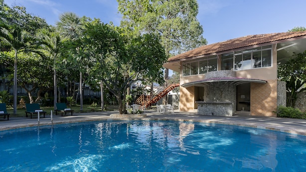 A swimming pool with a stone-tiled deck, surrounded by greenery and a building with a curved balcony.