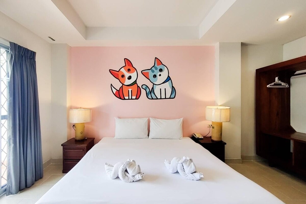 A hotel room with a large bed, two wall-mounted lamps, a window with curtains, and a wall decoration of two cartoon dogs.