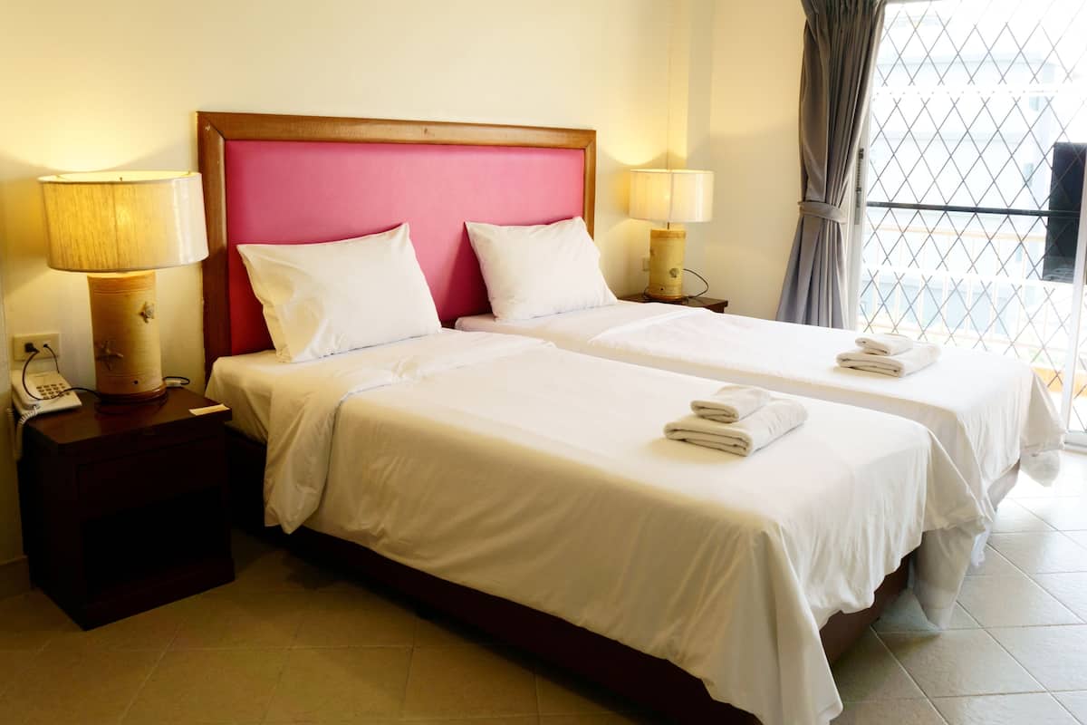 A hotel room with two beds, each with white linens and pillows, a wooden headboard, bedside lamps, and a window with a view of buildings.