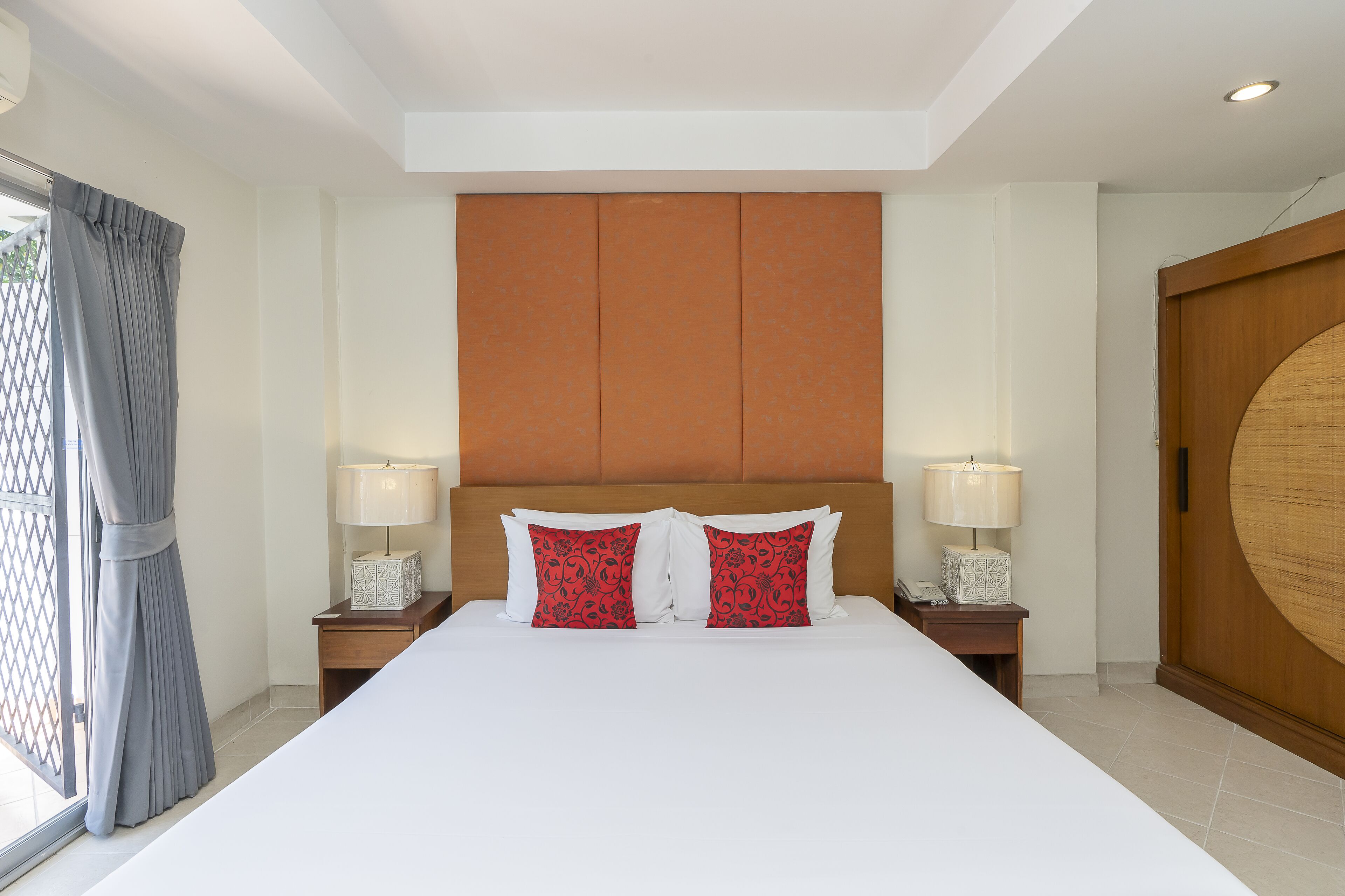A large bed with white linens and two red patterned pillows, flanked by nightstands with lamps, in a room with a wooden headboard and a large wooden door.