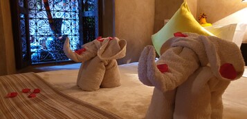 Un elefante de peluche y un conejo de peluche sobre una cama con pétalos de rosa, una ventana decorativa y una almohada amarilla.