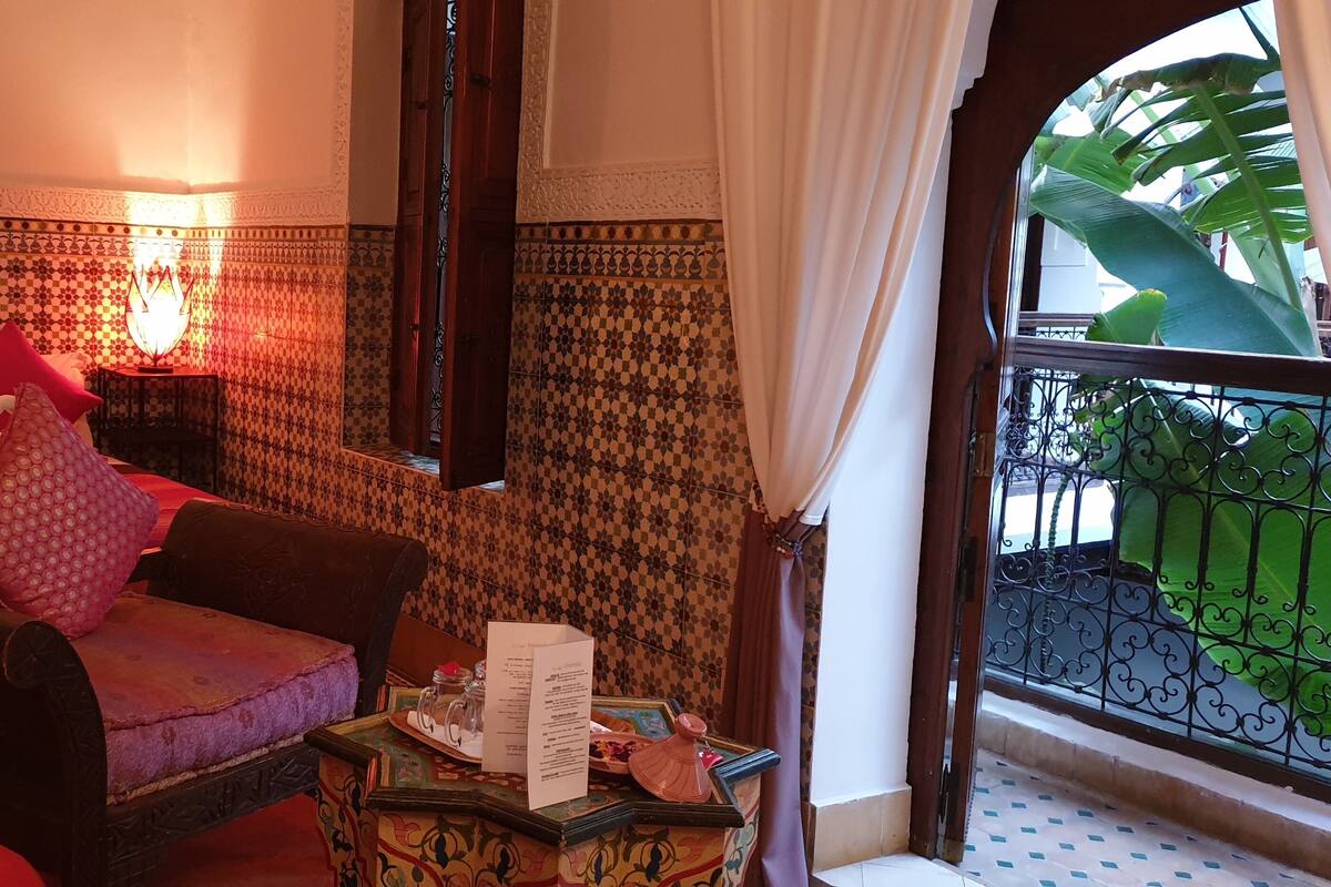 Habitación de hotel con cama, una silla, una mesa con un menú y vistas a un balcón con planta.