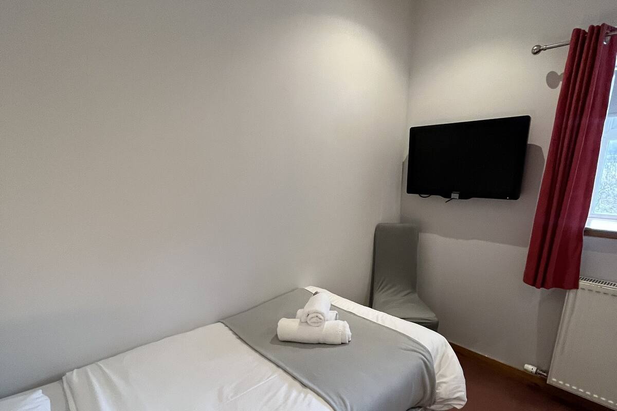 Un lit simple avec un matelas gris et du linge de lit blanc, un téléviseur à écran plat fixé au mur et une fenêtre avec des rideaux rouges.
