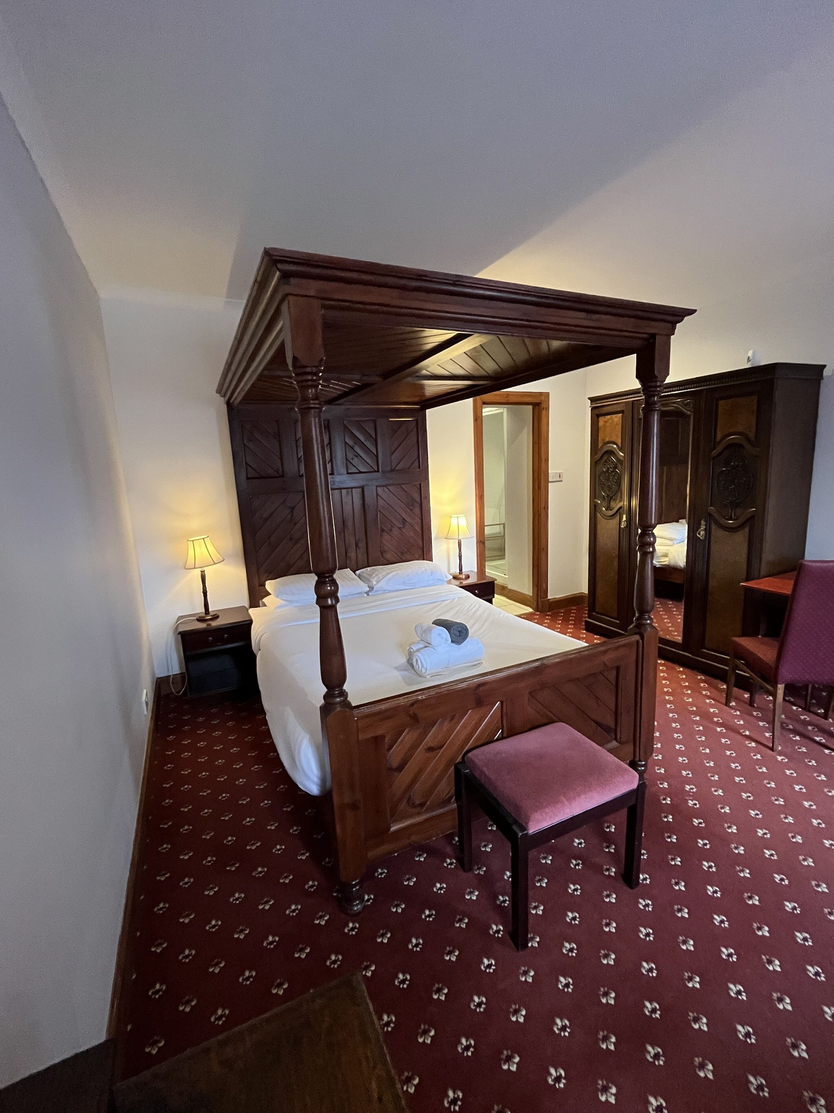 A four-poster bed with a wooden canopy, a bedside table with a lamp, a chair, and a wardrobe.