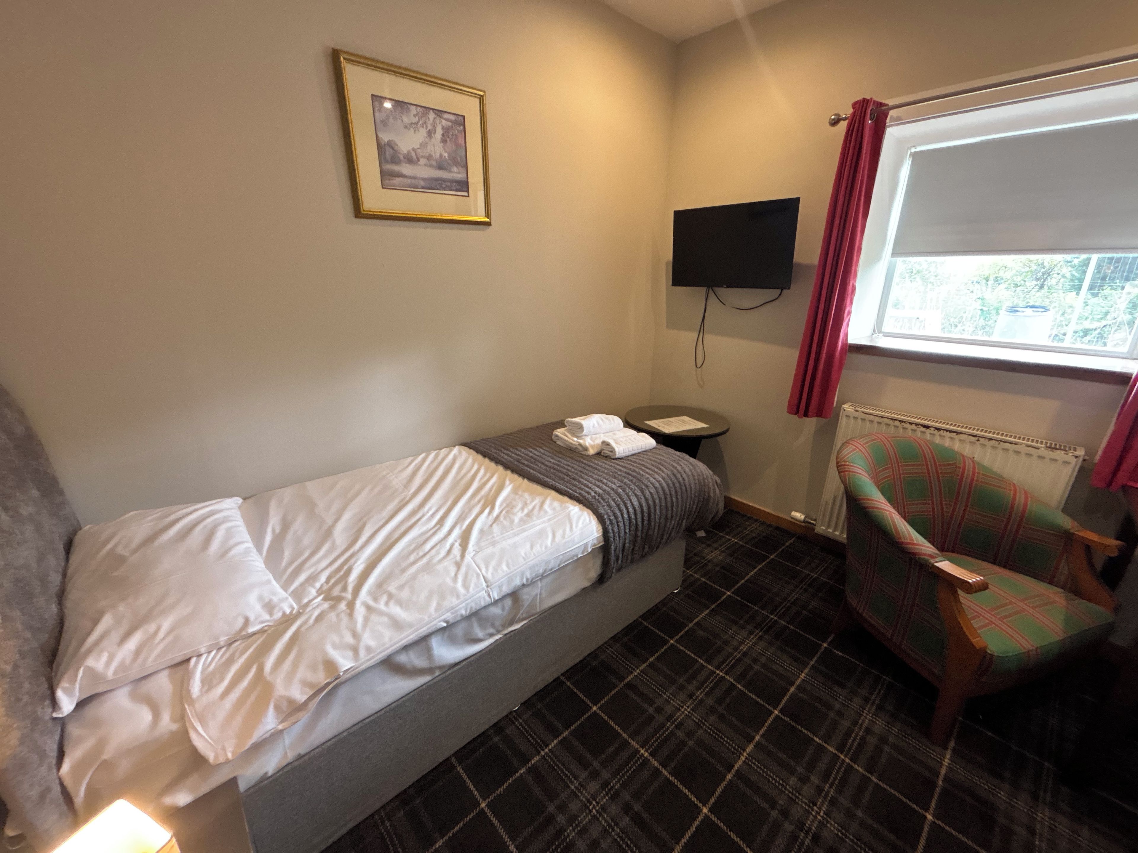 A single bed with white linens, a small bedside table with a lamp, a plaid-patterned chair, a wall-mounted TV, and a window with a view of greenery.