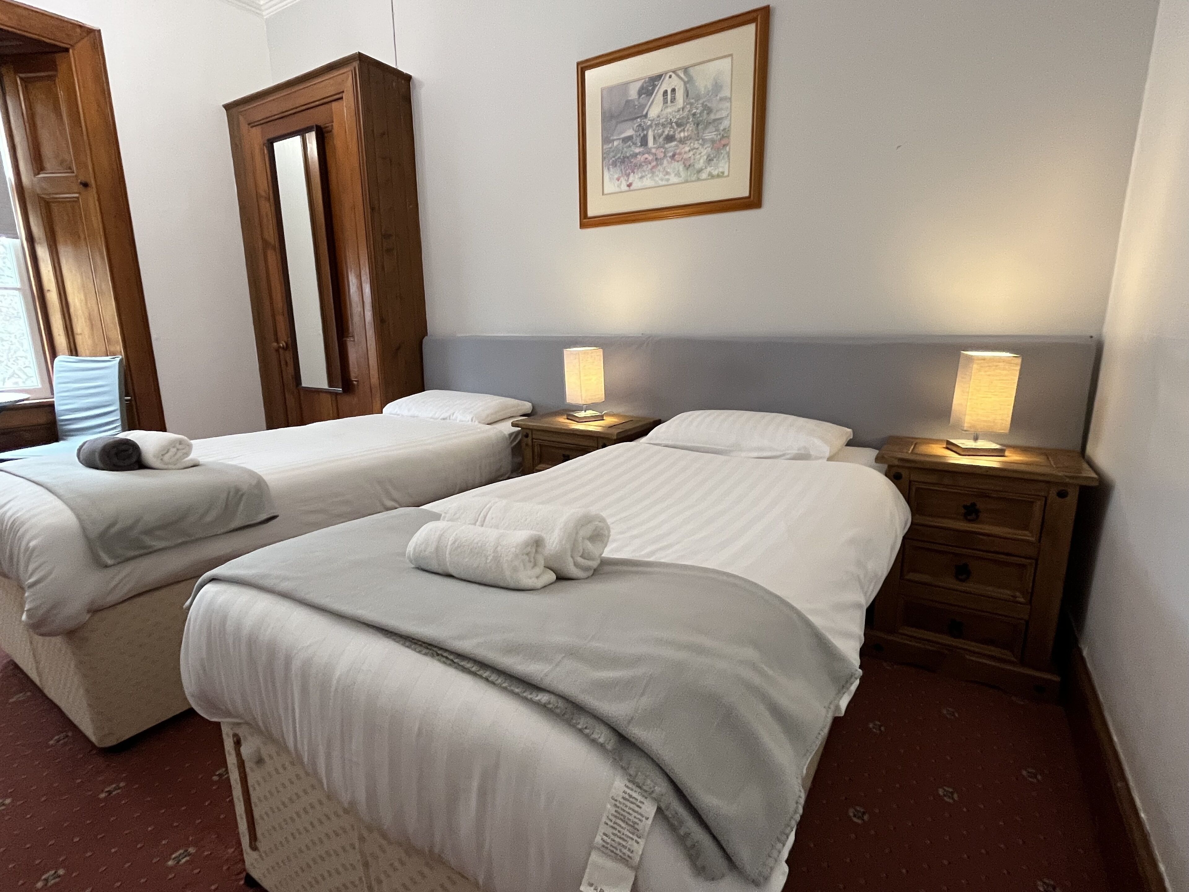 A hotel room with two beds, a wooden wardrobe, a framed picture on the wall, and bedside lamps.