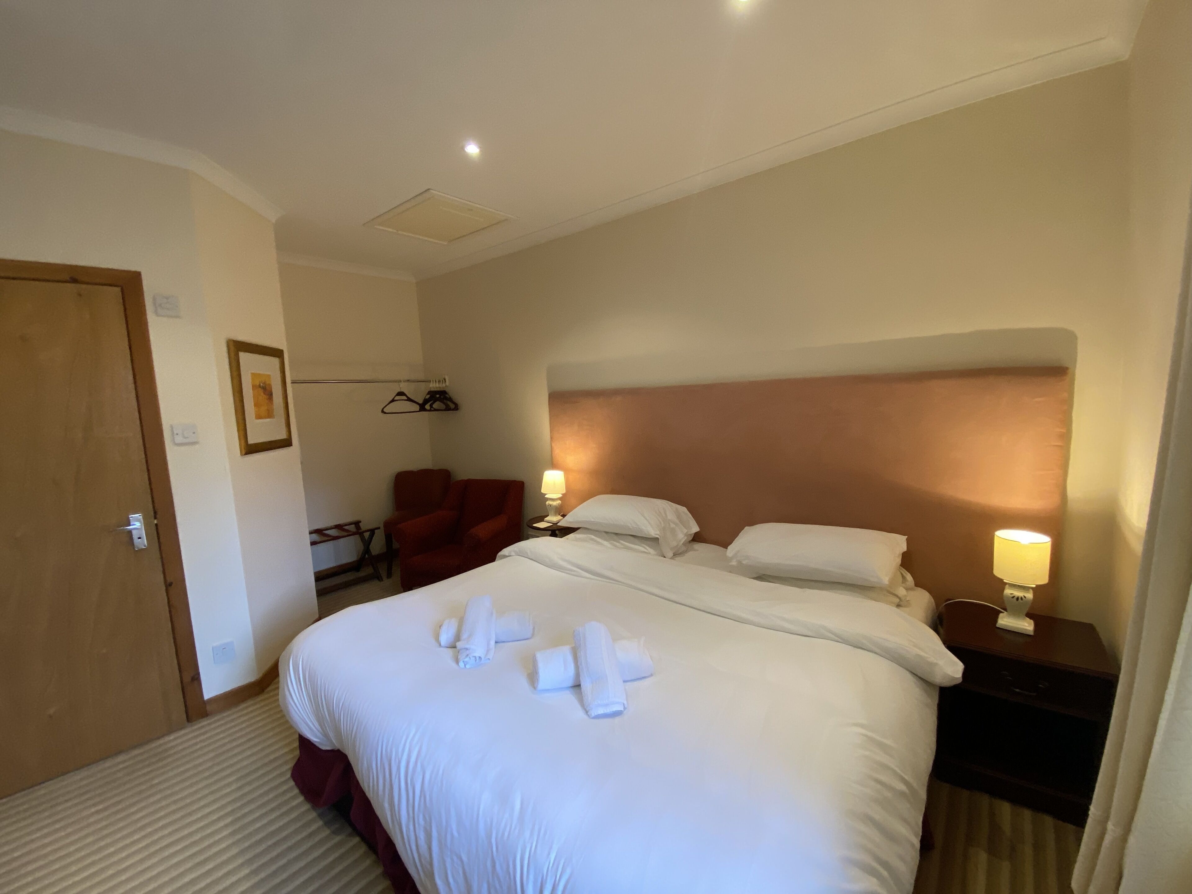 Una habitación de hotel con una cama grande, dos lámparas de noche, un sillón rojo y una mesita con lámpara.