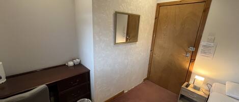 Et lille, godt oplyst soveværelse med en trædør, et skrivebord med stol, et natbord med lampe og en skraldespand.