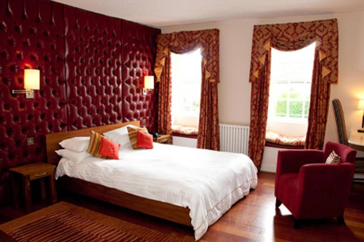 Ein Hotelzimmer mit einem großen Bett, einem roten Sessel und zwei mit Vorhängen versehenen Fenstern.