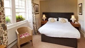 A bedroom with a large bed, two bedside tables with lamps, a wicker chair, and a window with curtains. - Woolpack Inn Beckington by Greene King Inns (Frome)