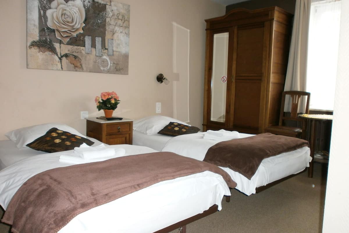 A hotel room with two beds, a wooden wardrobe, a mirror, a nightstand with a flower vase, and a painting on the wall.
