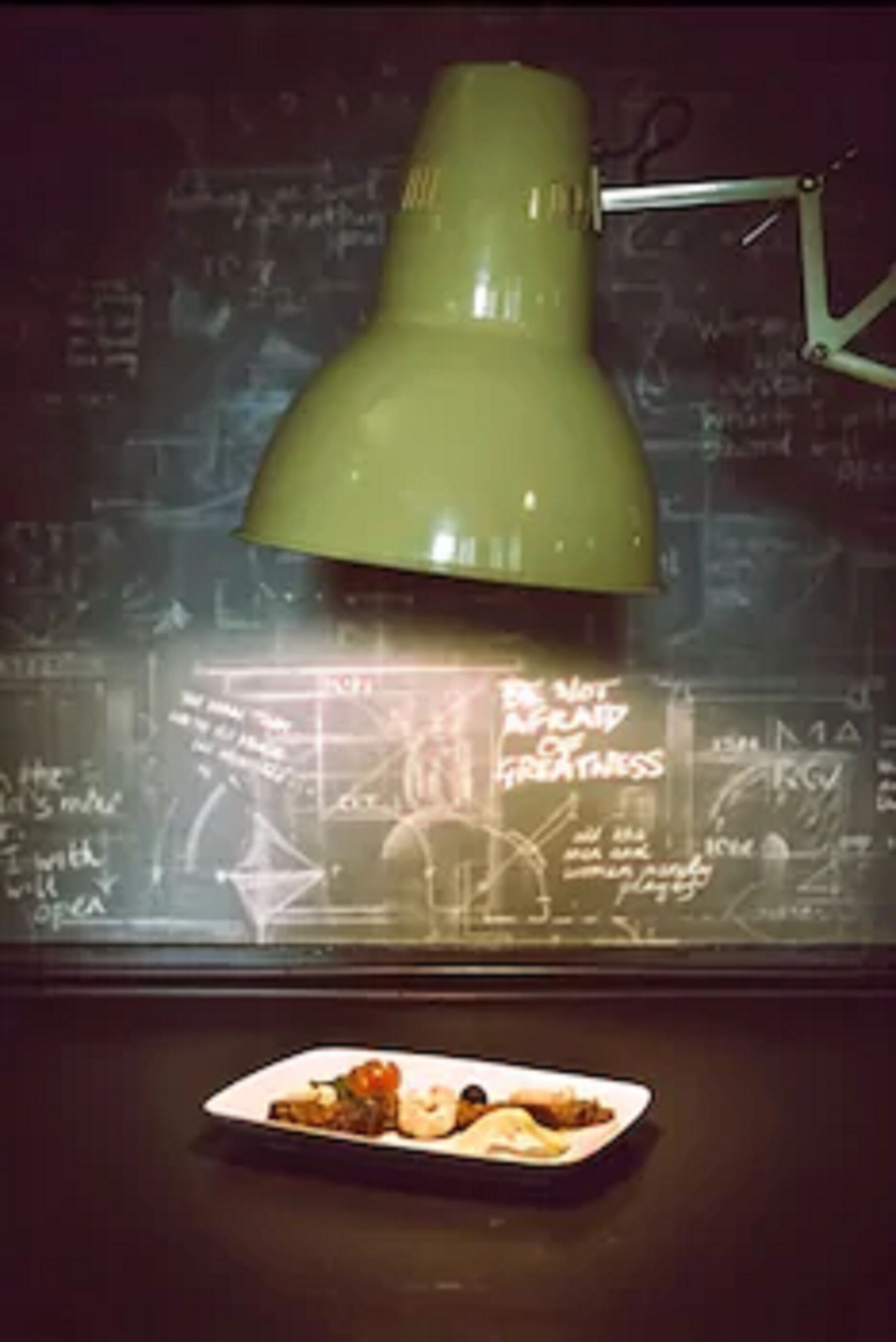 A table with a plate of food and a green desk lamp in front of a chalkboard with mathematical equations.