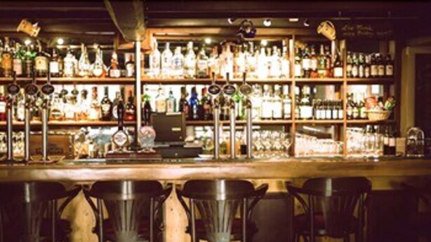 Un bar con múltiples estanterías llenas de diferentes botellas y vasos. Se ven varios taburetes en primer plano.