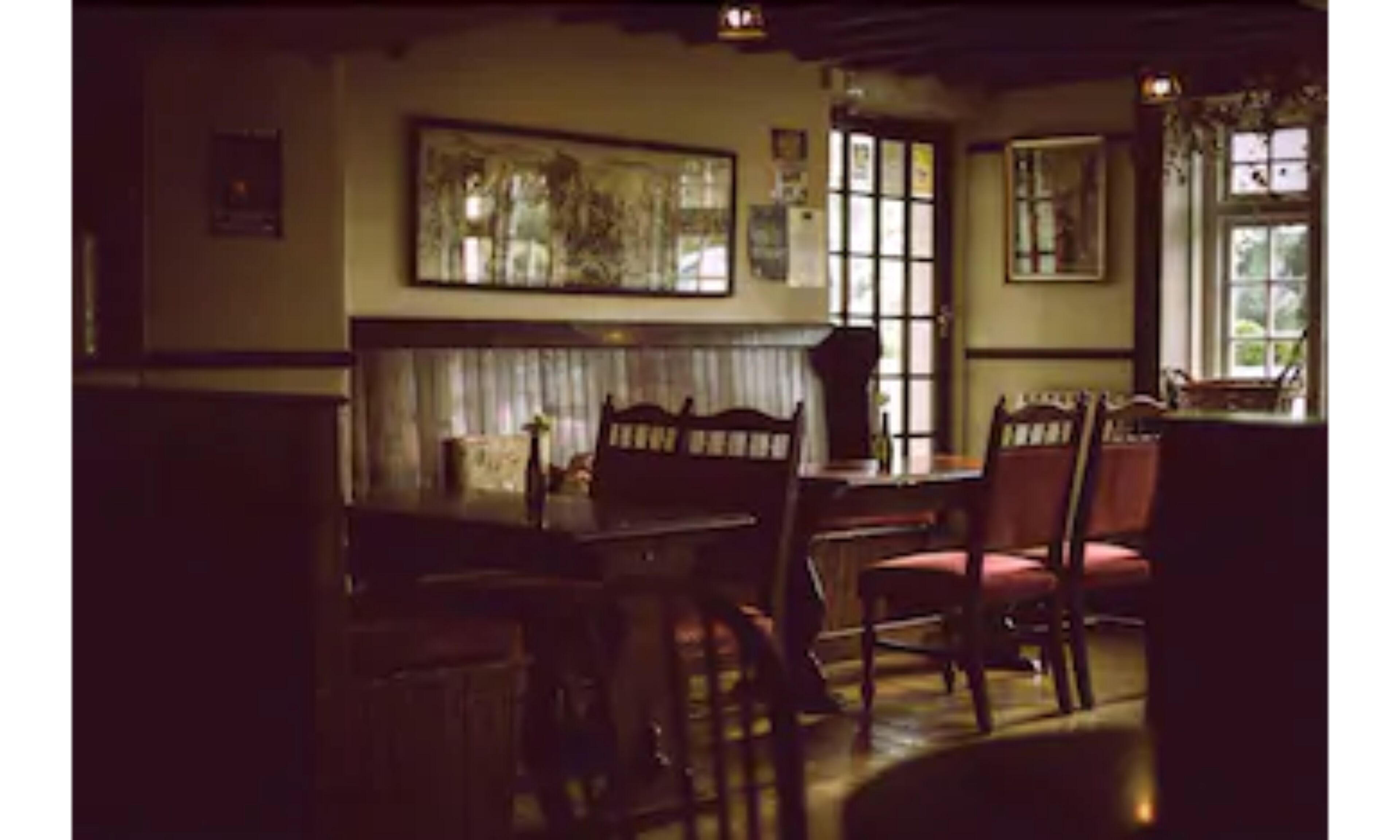 A dimly lit room with a wooden table and chairs, a large painting on the wall, and a window with multiple panes.