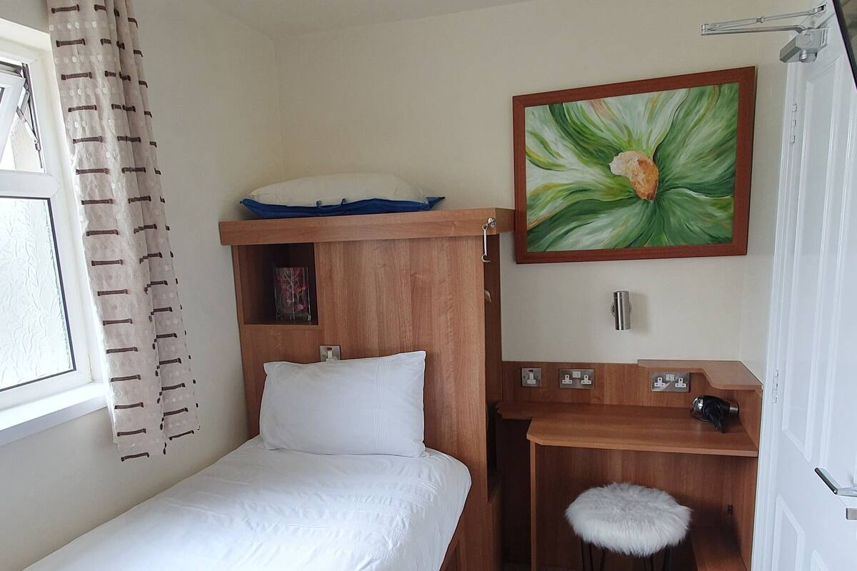 Ein kleiner, ordentlicher Raum mit einem Einzelbett, einem Schreibtisch und einem Stuhl. An der Wand hängt ein Gemälde, ein Fenster mit Vorhängen und eine Deckenleuchte.