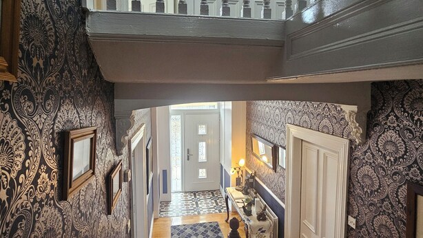 Una imponente escalera con diseños ornamentales en las paredes y un candelabro, que conduce a un pasillo luminoso con un alfombra estampada y una pequeña estatua.