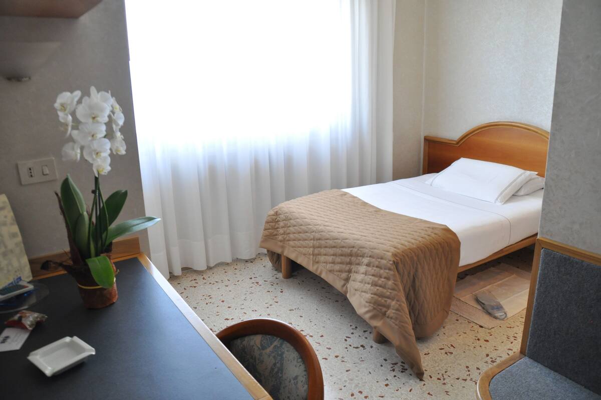 A hotel room with a bed, a desk, a chair, a potted orchid, and a window with curtains.
