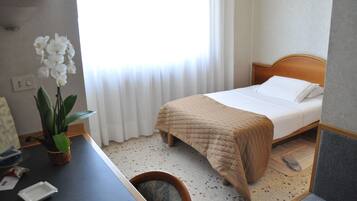 Una habitación de hotel con cama, escritorio, silla, orquídea en maceta y ventana con cortinas.