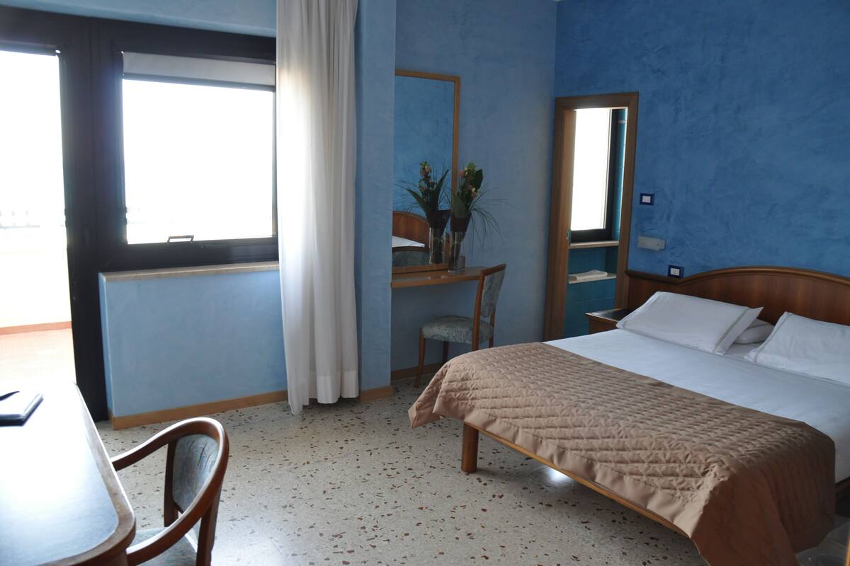 A hotel room with a bed, a desk, a chair, a mirror, and a window.