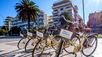 Une rangée de vélos est garée sur un trottoir, chacun muni d’un panier portant l’inscription « maja », et on aperçoit un panneau d’hôtel à l’arrière-plan.