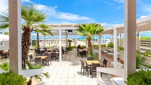 An outdoor patio area with tables and chairs, surrounded by palm trees, overlooking a beach. - Hotel Maja (Pescara)