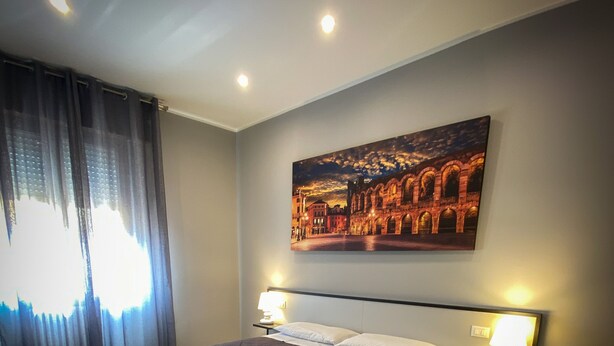 A modern bedroom with a large bed, two bedside tables with lamps, a ceiling light, and a cityscape painting on the wall. - Hotel Arena (Verona)
