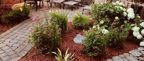 A patio area with stone and brick flooring, surrounded by various plants and shrubs, and includes outdoor furniture.